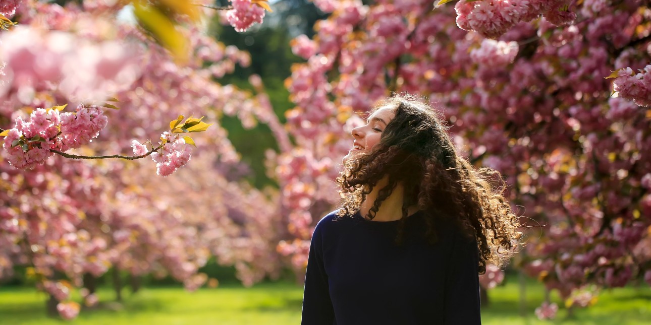 Young female with dark olive skin and long curly brown hair, smiling and enjoying the site of blooming cherry blossom trees. 