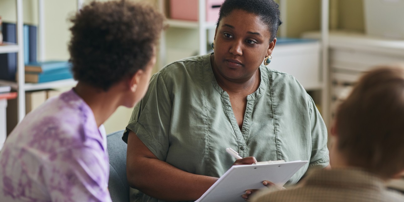 A Black woman takes notes and listens to a young man speaking.