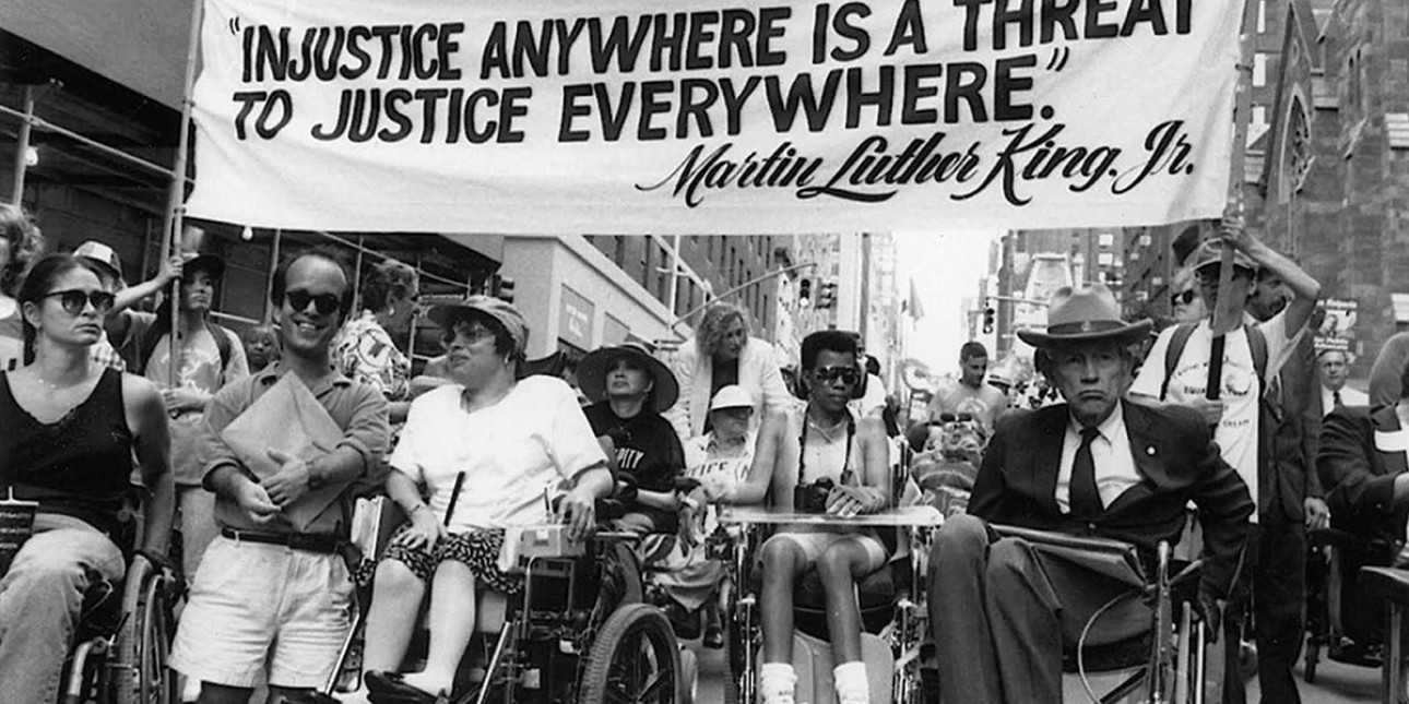 Black and white photo of several wheelchair-using participants in one of the first Disability Independence Day marches in New York City gathered under a banner that reads “‘Injustice Anywhere is a Threat to Justice Everywhere,’—Martin Luther King, Jr.” Jul 26, 1993