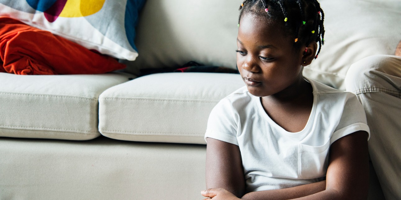 a Black girl sits in silence next to a couch