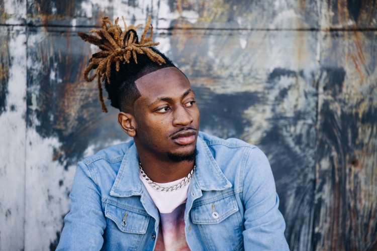 Yound adult male with brown skin wearing a white tshirt, silver chain necklace, and blue jean jacket sitting in front of a textured exterior wall.