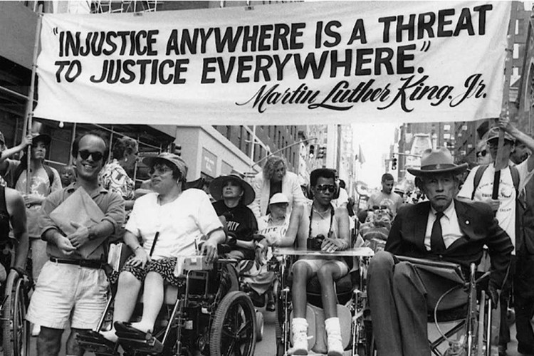 Black and white photo of several wheelchair-using participants in one of the first Disability Independence Day marches in New York City gathered under a banner that reads “‘Injustice Anywhere is a Threat to Justice Everywhere,’—Martin Luther King, Jr.” Jul 26, 1993