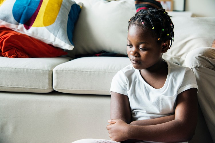 a Black girl sits in silence next to a couch